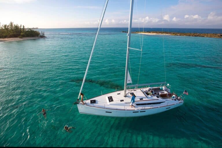 Ein weißer Katamaran liegt im klaren, türkisfarbenen Wasser in der Nähe einer kleinen, grünen Insel vor Anker. Zwei Personen schwimmen neben dem Boot, während eine andere Person auf dem Deck steht. Der Himmel ist größtenteils klar, mit einigen Wolken am Horizont. Es ist ein perfekter Tag für einen Segeltörn.