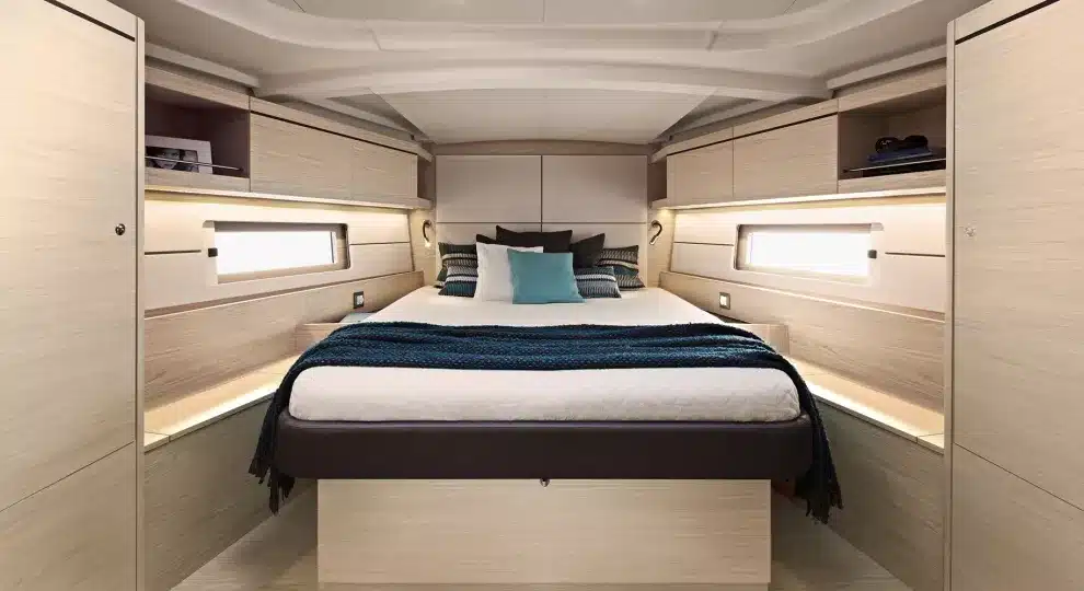 Ein kompaktes Schlafzimmer auf einer Katamaran-Yacht mit einem ordentlich gemachten Bett mit dunkelblauer und weißer Bettwäsche, umgeben von hellen Holzschränken und integrierten Regalen sowie dezenter Umgebungsbeleuchtung.