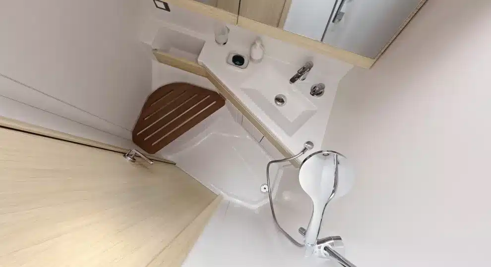 Ein kleines, modernes Badezimmer mit strahlend weißen Wänden und Boden, mit einer Eckdusche, einem Waschbecken mit Chromarmaturen, einem großen Spiegel und einem Toilettensitzdeckel aus Holz.