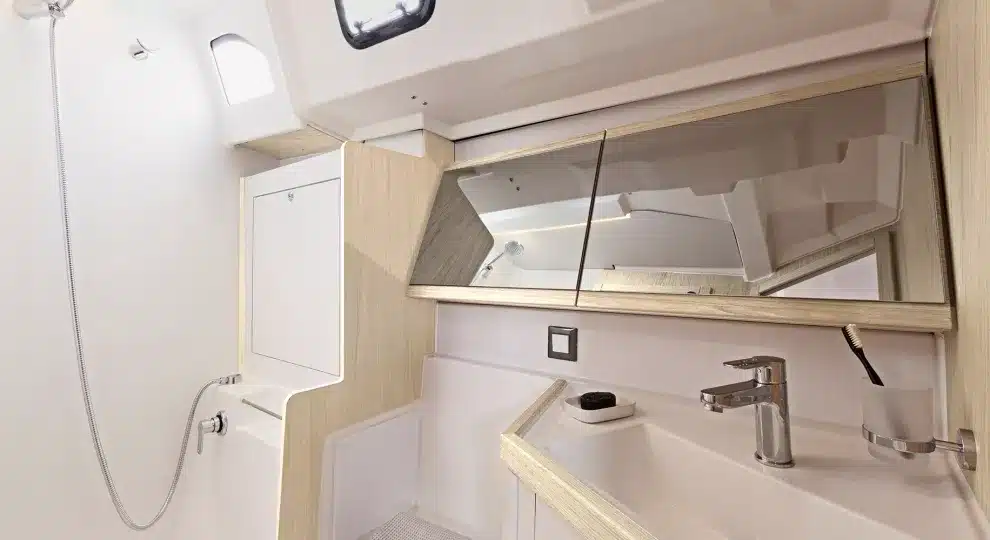 Kompaktes Bootsbadezimmer mit weißen Wänden, einem kleinen Waschbecken, einem Spiegel und einem Duschbereich auf der linken Seite. Helle Holzpaneele ergänzen das moderne, minimalistische Katamaran-Design.