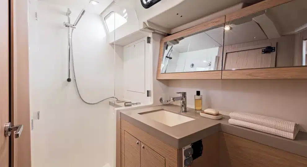 Ein modernes, kompaktes Segelyacht-Badezimmer mit einer wandmontierten Dusche, Holzschränken, einem rechteckigen Waschbecken, einem Spiegel und gut sortierten Handtüchern unter einer weißen Decke mit Oberlicht.