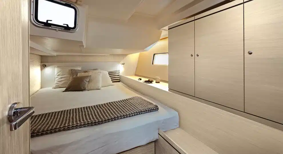 Innenansicht einer Luxusyachtkabine mit ordentlich gemachtem Bett, Holzschränken und sanfter Beleuchtung sowie einem kleinen Fenster an der Seite während eines Segeltörns.
