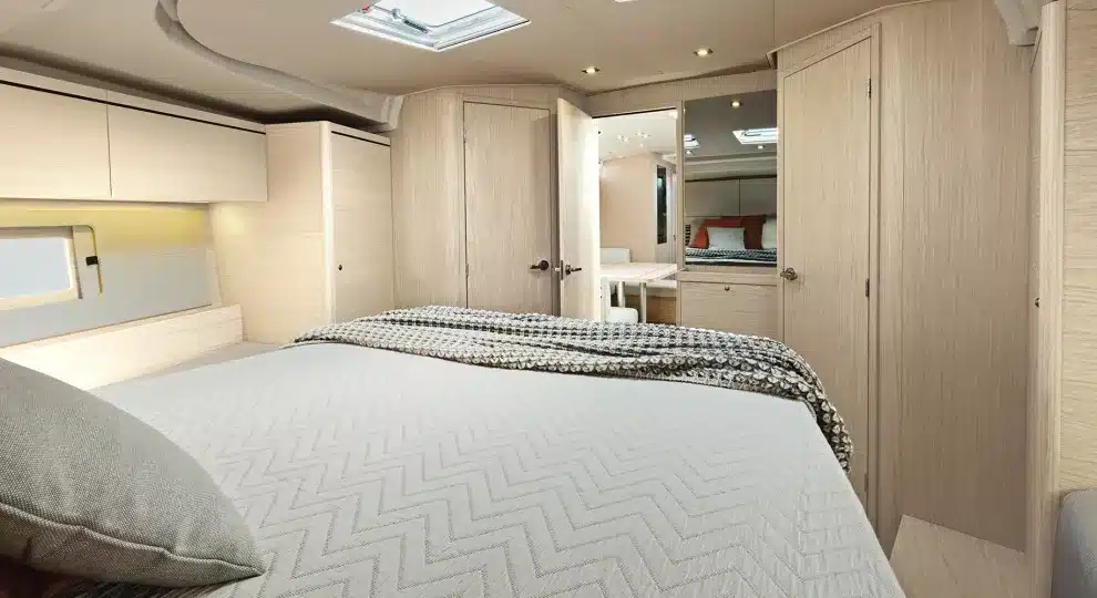 Innenansicht einer modernen Segelyachtkabine mit einem großen Bett mit einer grau-weiß gemusterten Bettdecke, umgeben von Holzschränken und natürlichem Licht, das durch die Fenster strömt.