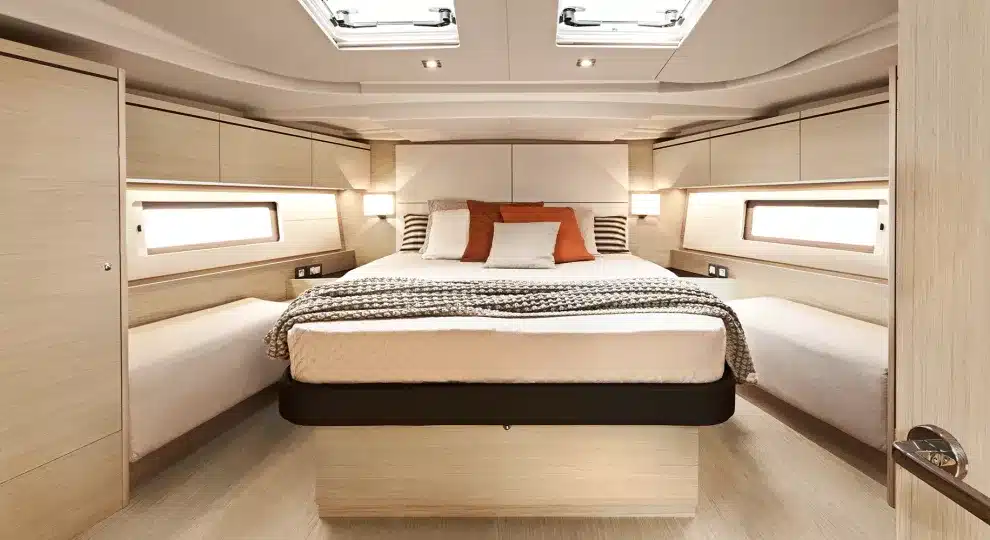 Luxuriöses Segelyacht-Schlafzimmer mit großem Bett mit weißer und orangefarbener Bettwäsche, umgeben von Holzpaneelen und Schränken. Zwei Bullaugenfenster sorgen für Tageslicht.