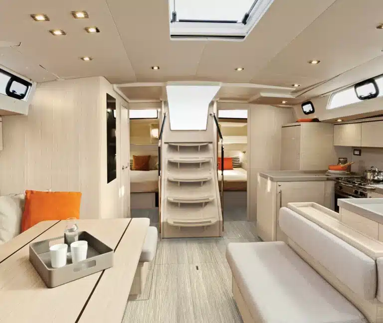 Innenansicht einer modernen Segelyacht mit einem gemütlichen Loungebereich mit orangefarbenen Kissen, einer integrierten Küche und einer Treppe, die zu einem anderen Deck führt, alles mit natürlichem Licht von oben.