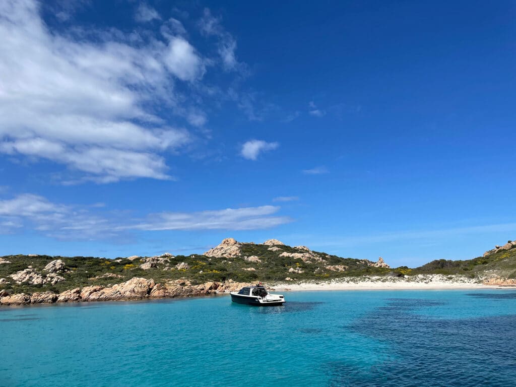 Ein kleines Motorboot schwimmt auf klarem türkisfarbenem Wasser in der Nähe einer felsigen Küste unter einem blauen Himmel mit vereinzelten weißen Wolken. Im Hintergrund erheben sich schroffe, mit Sträuchern bewachsene Hügel, an denen eine anmutige Segelyacht vorbeigleitet und die ruhige und malerische Küstenlandschaft noch verstärkt.