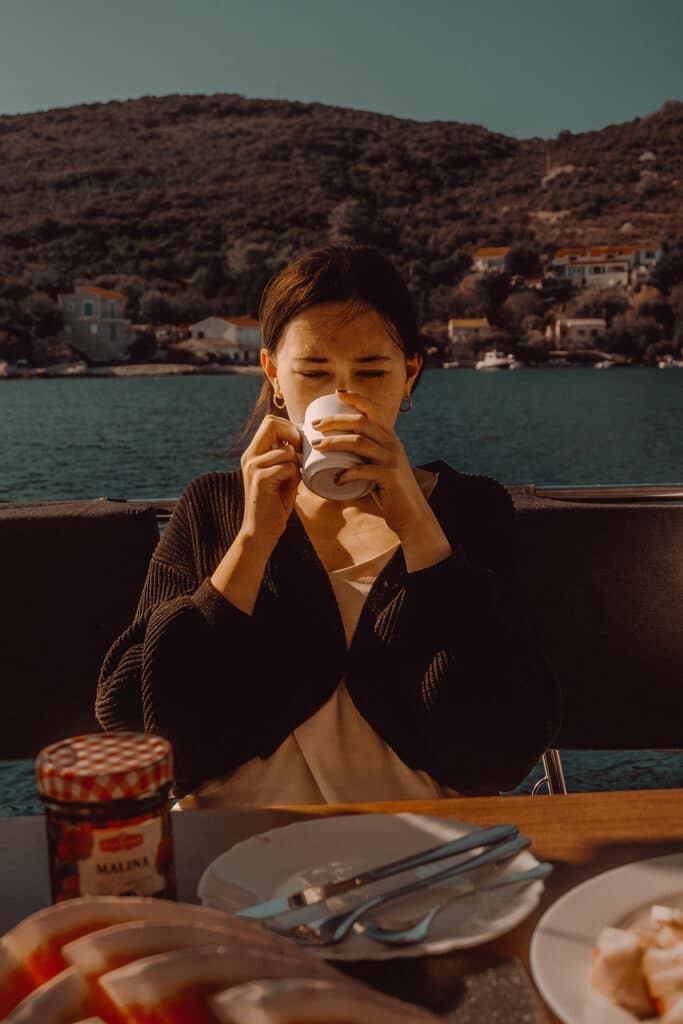 Eine Person sitzt auf einem Boot, nippt mit geschlossenen Augen an einer Tasse und genießt einen friedlichen Moment. Im Hintergrund sind ein ruhiges Gewässer und hügeliges Gelände mit Häusern zu sehen. Im Vordergrund auf dem Tisch stehen ein Glas Marmelade und ein Teller mit Essen während ihres ruhigen Segelurlaubs.