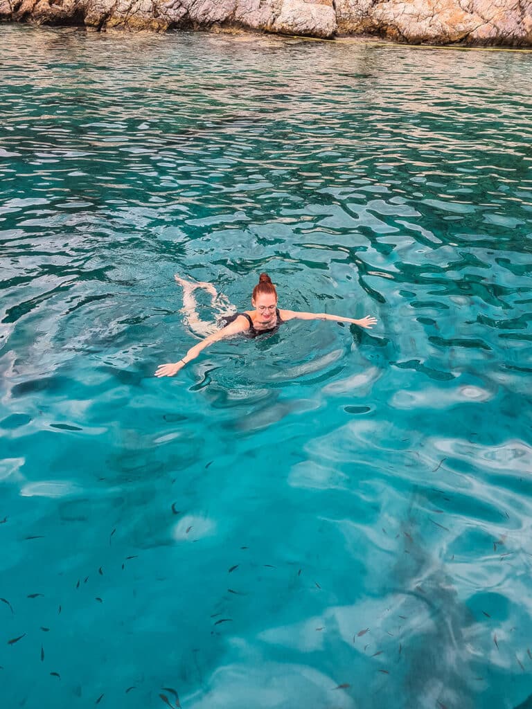Eine Person schwimmt im klaren, türkisfarbenen Wasser. Kleine Fische schwimmen in der Nähe. Mit ausgestreckten Armen scheint sie das erfrischende Wasser zu genießen. Im Hintergrund sind ein felsiges Ufer und eine Segelyacht von ihrer Segelreise zu sehen.