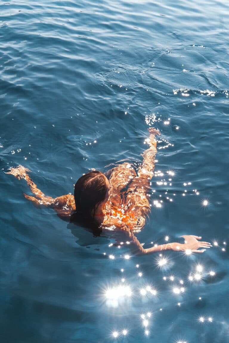 Eine Person mit langen Haaren schwimmt in klarem, blauem Wasser. Das Sonnenlicht spiegelt sich auf der Wasseroberfläche und erzeugt glitzernde Muster um sie herum. Die Szene ist ruhig, und als sie in der Nähe eines Katamarans dahintreibt, ist klar, dass sie einen friedlichen Moment während ihres Segelurlaubs genießt.