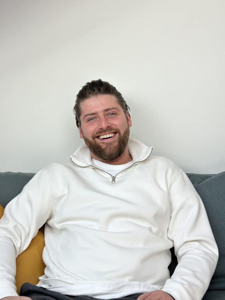 Ein Mann mit kurzen braunen Haaren und Bart sitzt lächelnd auf einem Sofa. Er trägt einen weißen Pullover mit Kragen, der an die Pullover erinnert, die man im Segelurlaub trägt. Der Hintergrund ist eine schlichte weiße Wand und neben ihm liegt ein senfgelbes Kissen.