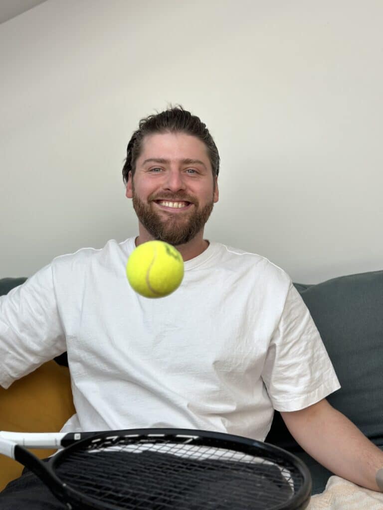 Ein Mann mit Bart und weißem T-Shirt sitzt auf einem Sofa und lächelt. Er hält einen Tennisschläger und vor ihm schwebt ein Tennisball in der Luft. Er träumt von seiner nächsten Segelreise. Der Hintergrund ist neutral mit einer grauen Wand und Kissen auf dem Sofa.