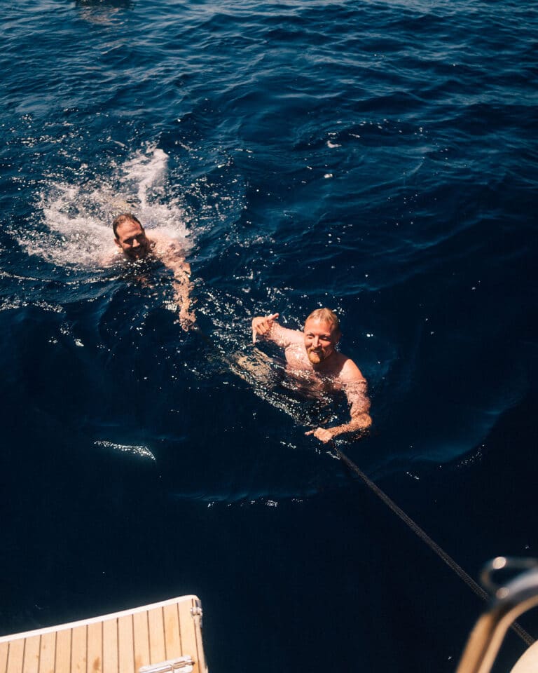Zwei Menschen schwimmen während ihres Segelurlaubs im tiefblauen Meerwasser. Einer planscht in der Nähe, während der andere, näher an einer Holzplattform, sich an einem Seil festhält und lächelnd nach oben schaut. Die Szene wirkt lebendig und fröhlich mit sonnenbeschienenen Spiegelungen auf dem Wasser.