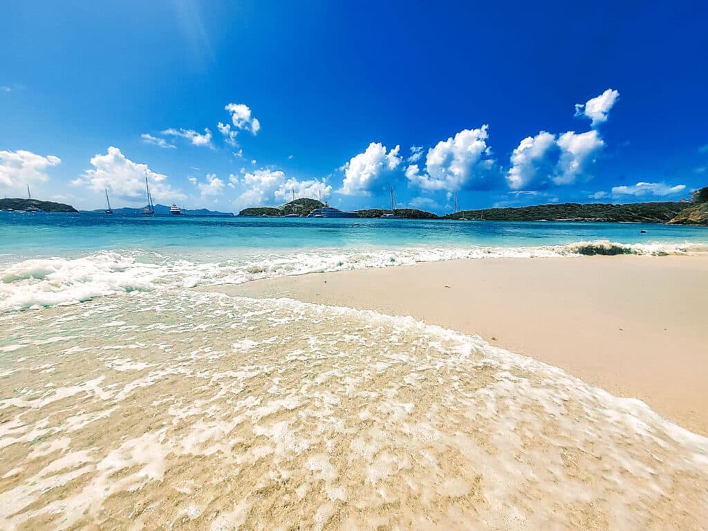 Eine ruhige Strandszene mit sanften Wellen, die unter einem strahlend blauen Himmel an das Sandufer plätschern. Im Hintergrund ankern Katamarane und Segelboote in der Nähe üppiger grüner Hügel. Flauschige weiße Wolken sind über den Himmel verstreut und verstärken die idyllische Küstenatmosphäre.