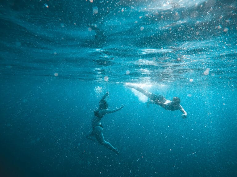 Zwei Menschen schwimmen unter Wasser in einem klaren, blauen Ozean neben einer majestätischen Segelyacht. Die Person auf der linken Seite streckt ihre Arme mit einer anmutigen Schwimmbewegung aus, während die Person auf der rechten Seite tritt, um sich vorwärts zu bewegen. Sonnenlicht fällt durch und erzeugt einen faszinierenden, schimmernden Effekt im Wasser.