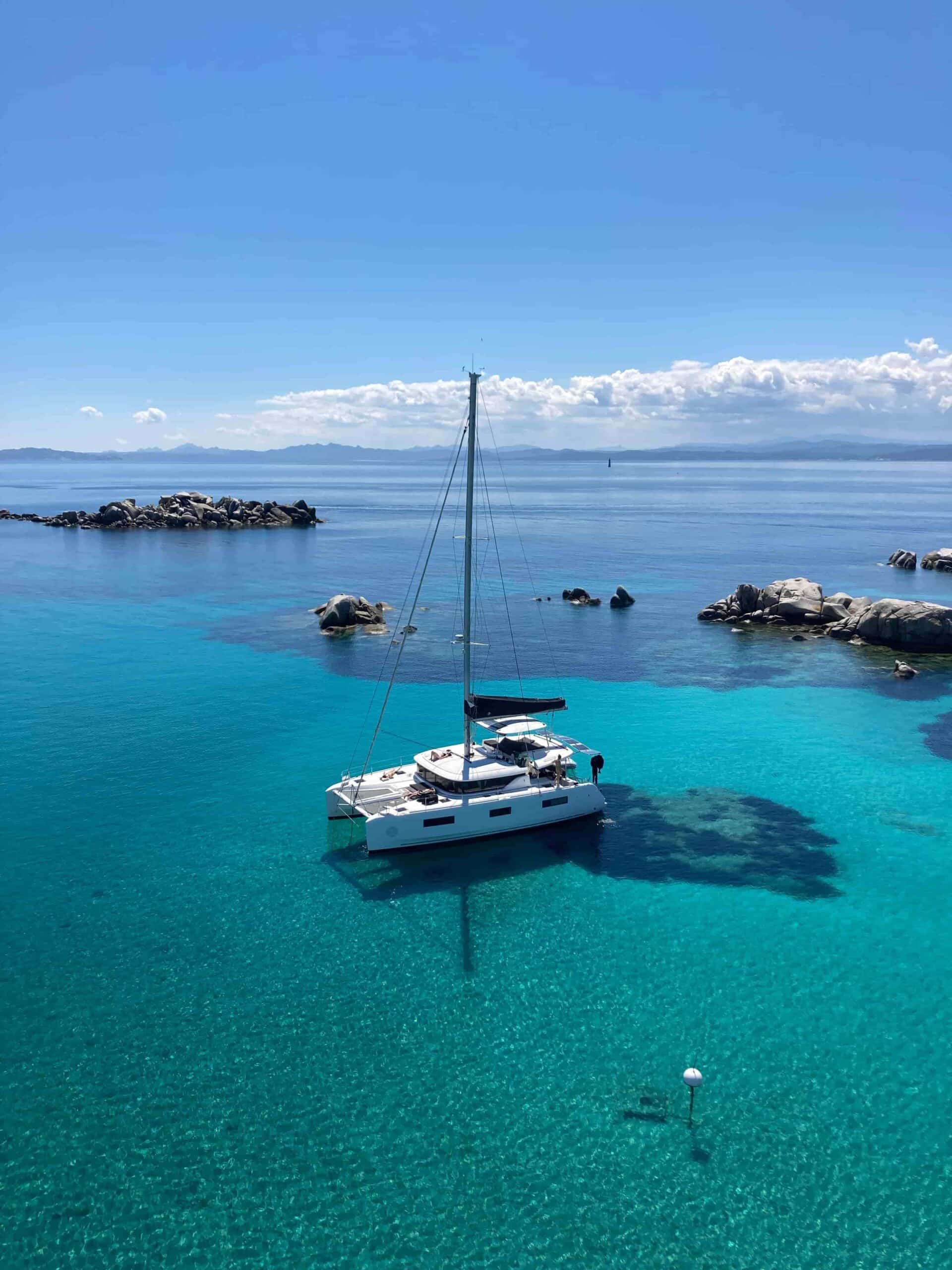Ein weißes Segelboot ankert in klarem, türkisfarbenem Wasser mit einer felsigen Küste im Hintergrund. Das ruhige Meer spiegelt den blauen Himmel mit flauschigen Wolken und schafft eine ruhige und idyllische Szene, perfekt für einen Segeltörn.