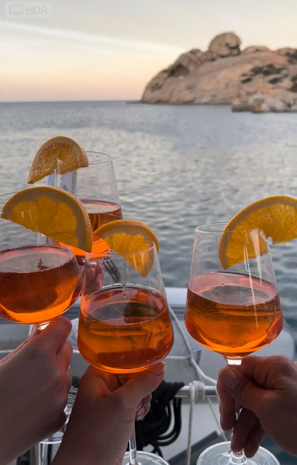 Vier Hände heben Gläser mit einem Orangengetränk, garniert mit Orangenscheiben, vor einem malerischen Blick auf ruhiges Wasser und felsige Klippen bei Sonnenuntergang. Die festliche Stimmung lässt auf eine Feier in Küstennähe schließen, vielleicht nach einem aufregenden Segeltörn auf einer luxuriösen Segelyacht.
