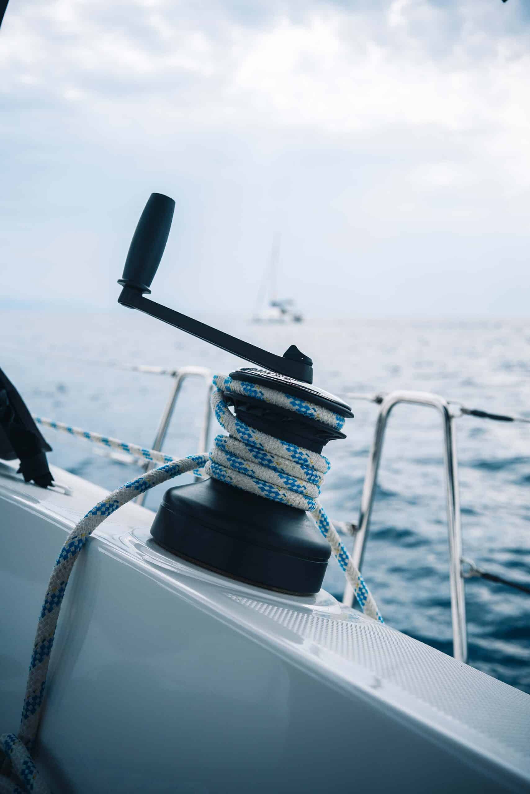 Nahaufnahme einer Winde auf einem Segelboot während eines Segelurlaubs, um die ein blau-weißes Seil gewickelt ist. Das Boot liegt auf dem Wasser, darüber ein bewölkter Himmel. Die Horizontlinie und ein weiteres Segelboot in der Ferne sind sichtbar und fangen die Essenz eines ruhigen Segeltörns ein.