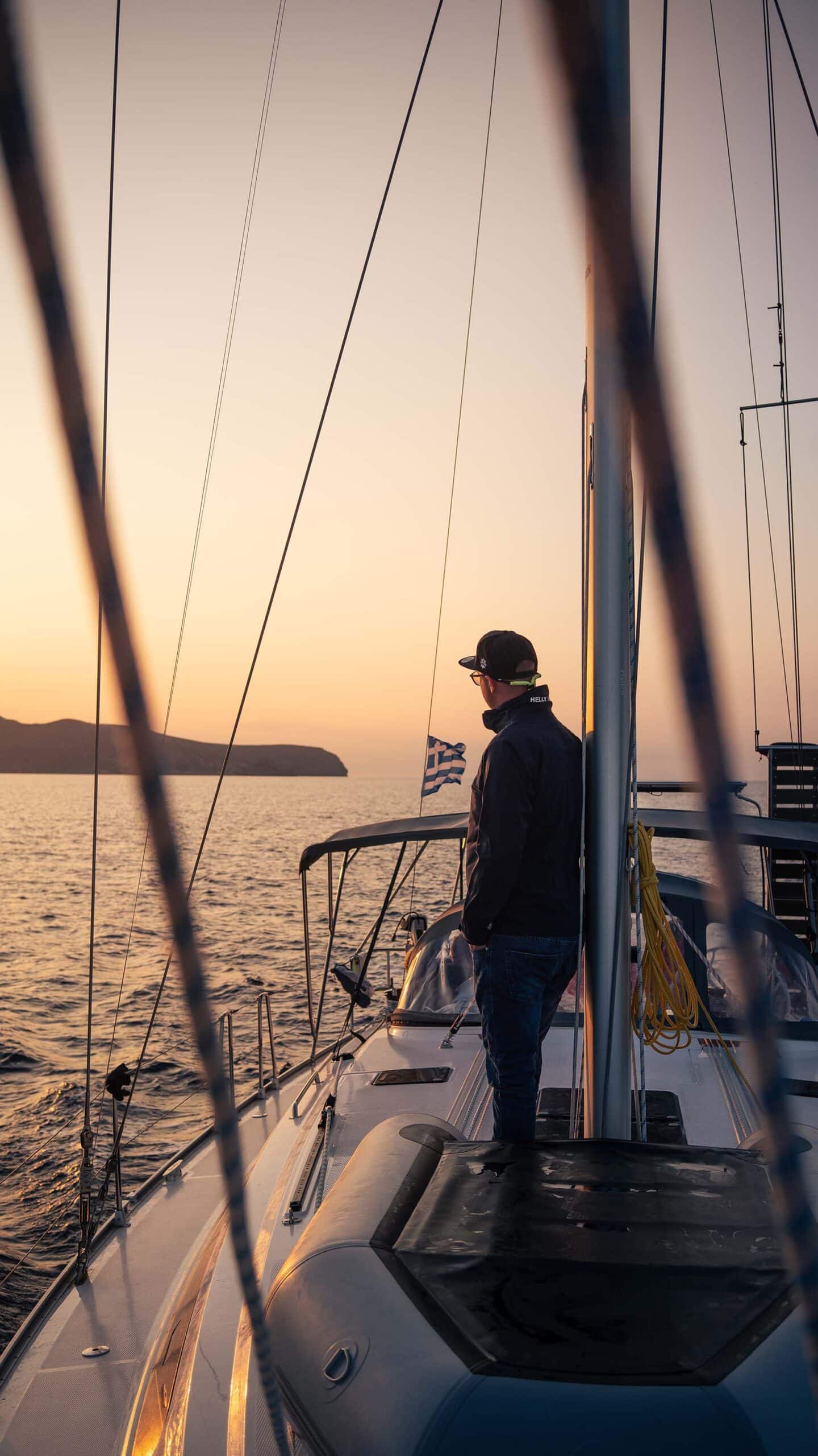Eine Person steht bei Sonnenuntergang auf dem Deck eines Segelboots, genießt ihren Segeltörn und blickt zum Horizont. Im Vordergrund sind die Takelage und Seile des Segelboots zu sehen, im Hintergrund ist eine Insel über einem ruhigen Meer zu sehen. Die warmen Farben der untergehenden Sonne tauchen diese idyllische Segelurlaubsszene in einen ruhigen Schein.