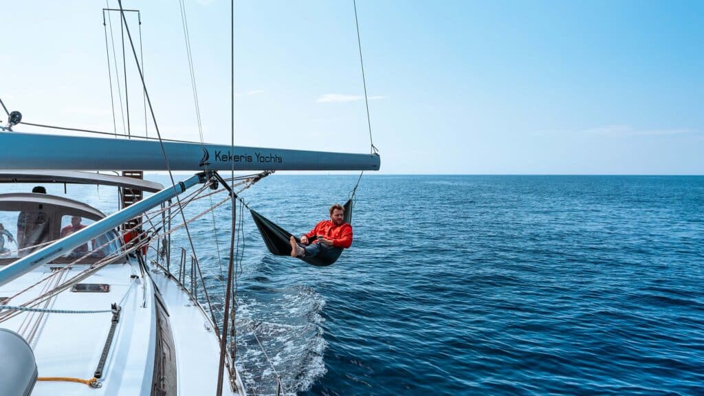 Eine Person in roter Jacke und schwarzen Shorts entspannt sich in einer Hängematte, die am Baum einer Segelyacht mit dem Aufdruck „Kekeris Yachts“ hängt. Das Boot segelt auf einem ruhigen blauen Meer unter klarem Himmel und genießt den ultimativen Segeltörn.