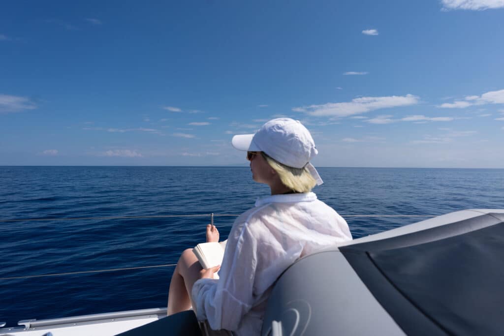 Eine Person mit weißem Hut, Sonnenbrille und weißem Hemd sitzt am Rand einer Segelyacht und liest ein Buch. Vor ihr erstreckt sich das Meer unter einem klaren blauen Himmel mit ein paar Wolken. Meer und Himmel dominieren die ruhige Szene während dieses heiteren Segelurlaubs.