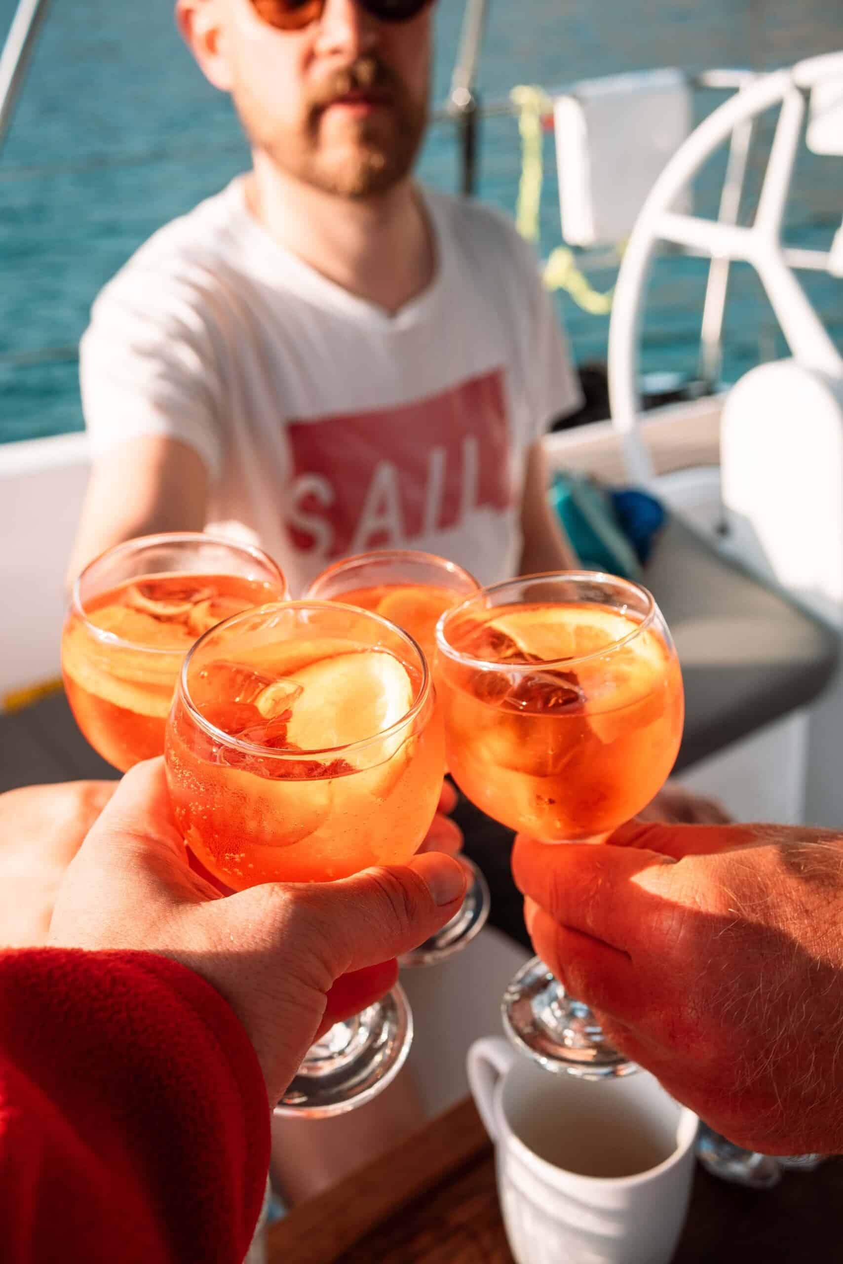 Vier Personen stoßen auf einer Segelyacht mit einem orangefarbenen Getränk an. Der Fokus liegt auf den Getränken im Vordergrund, im Hintergrund ist eine Person mit Sonnenbrille und „SAIL“-T-Shirt zu sehen. Die Szene wirkt entspannt und feierlich und fängt die Freude am Mitsegeln ein.
