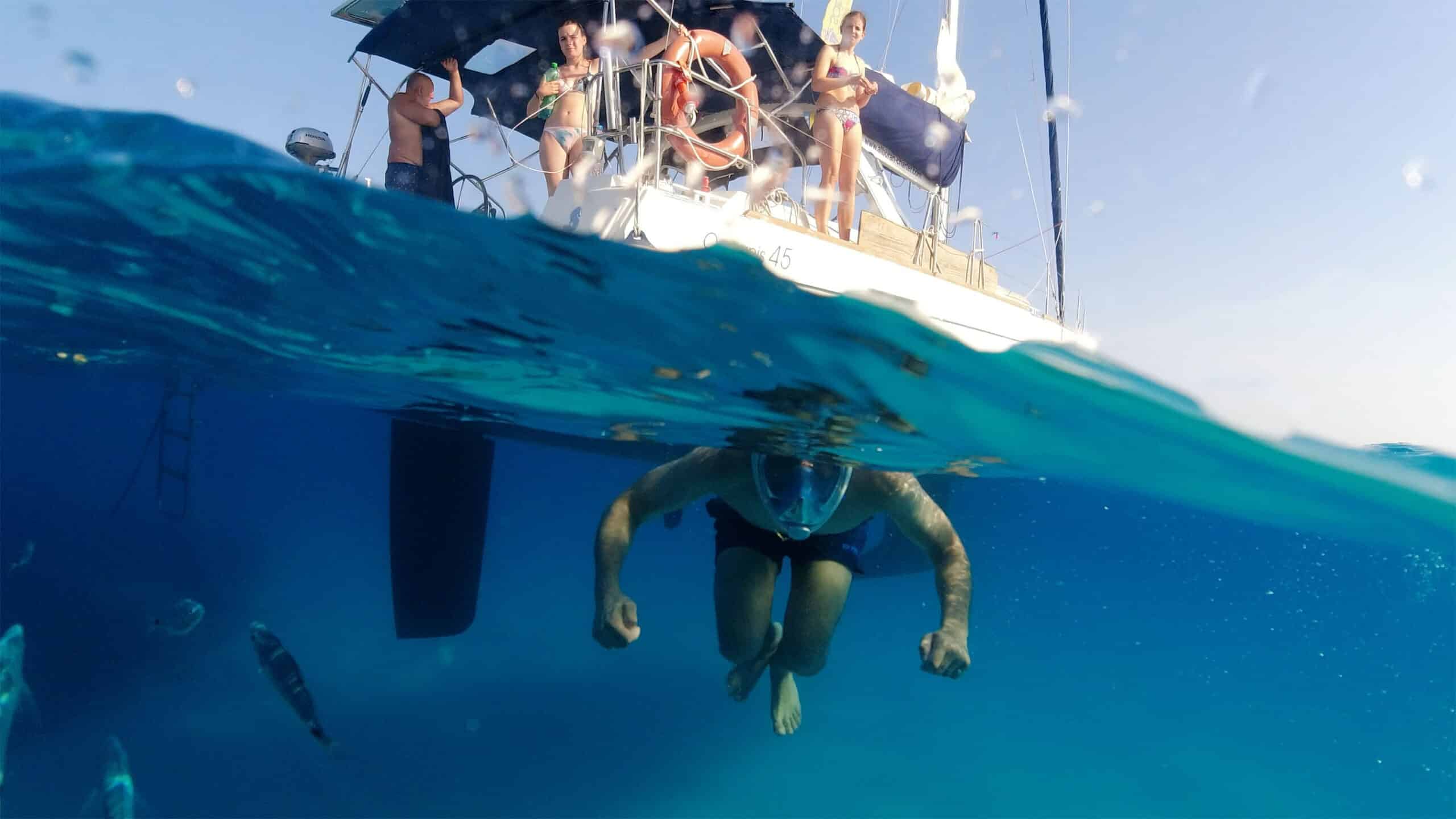 Ein Schnorchler schwimmt unter Wasser neben einem schnittigen Katamaran mit Menschen an Bord. Das Bild fängt sowohl die Unterwasserszene ein, auf der ein paar Fische zu sehen sind, als auch das Boot darüber, auf dem mehrere Menschen in Badebekleidung zu sehen sind, die auf den Schwimmer herabblicken.