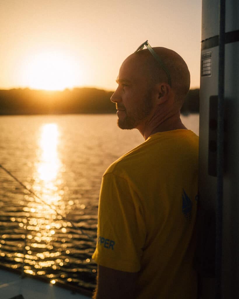 Ein Mann in einem gelben Hemd steht bei Sonnenuntergang auf einem Boot und blickt aufs Wasser. Er trägt eine Sonnenbrille auf dem Kopf und das sanfte goldene Licht der untergehenden Sonne spiegelt sich im Wasser und schafft eine heitere Atmosphäre – perfekt, um über einen Langzeiturlaub nachzudenken.