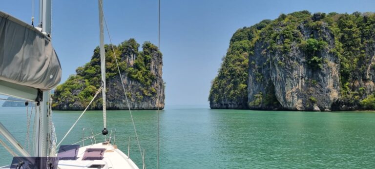 Eine unberührte Meereslandschaft mit dem Bug einer eleganten Segelyacht, die auf zwei hoch aufragende, felsige Inseln zusteuert, die von üppigem Grün bedeckt sind. Das ruhige türkisfarbene Wasser bildet einen Kontrast zum klaren blauen Himmel und schafft eine ruhige und malerische Kulisse, die perfekt für eine unvergessliche Segelreise ist.