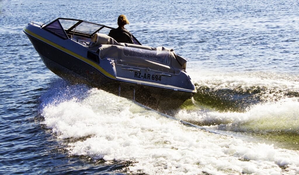 Eine Person mit langen Haaren, die stolz ihr SBF See zur Schau stellt, steuert an einem sonnigen Tag ein Schnellboot mit dem Kennzeichen „RZ-AR 694“. Das Boot hinterlässt eine Spur weißer Wellen im blauen Wasser.