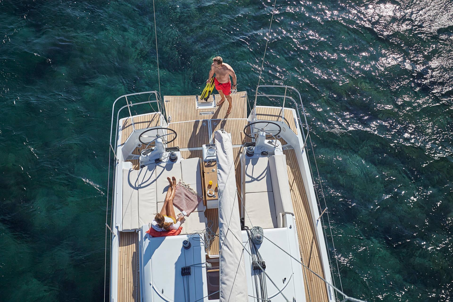 Luftaufnahme eines Segelboots auf klarem, grünem Wasser. Zwei Personen sind an Bord; eine breitet ein Handtuch auf dem Deck aus und trägt einen Hut, während die andere am Heck steht, gelbe Flossen hält und eine rote Badehose trägt – eine idyllische Szene, perfekt für Yachtcharter-Enthusiasten.