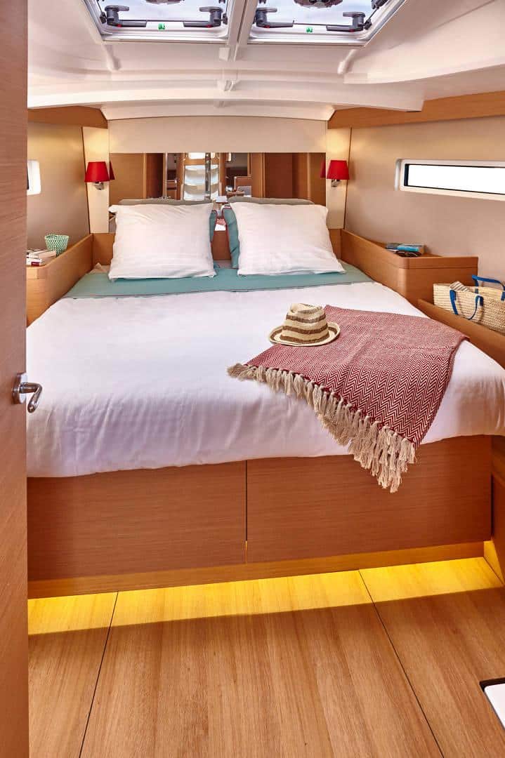 Ein gemütliches Schlafzimmer auf einem Yachtcharterboot verfügt über ein Doppelbett mit weißer Bettwäsche, einer roten Decke und einem darauf liegenden Hut. An den holzgetäfelten Wänden sind zwei Leselampen mit roten Schirmen angebracht, während natürliches Licht durch ein Fenster einfällt.