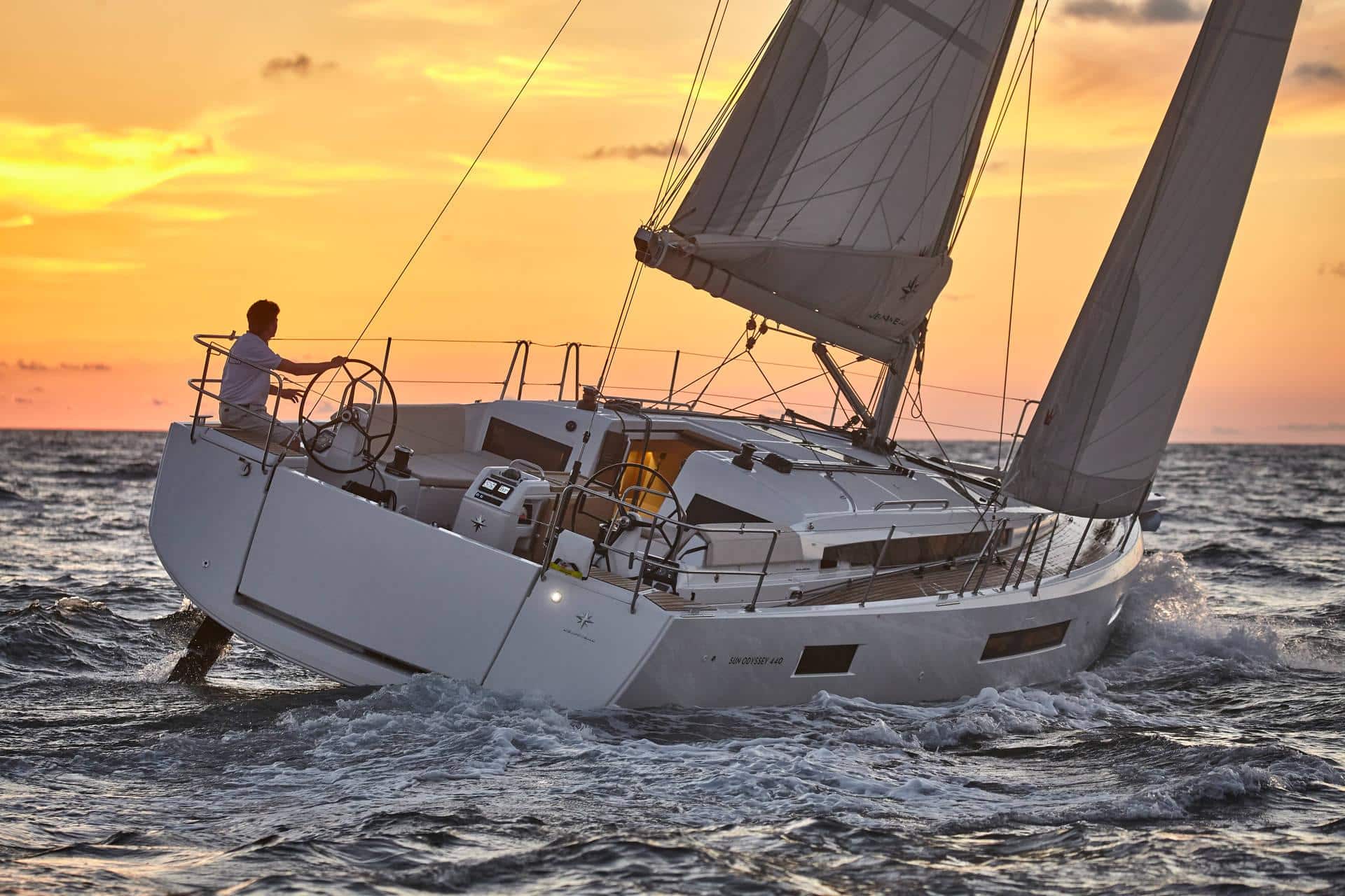 Ein einsamer Segler steuert ein weißes Segelboot bei Sonnenuntergang auf rauer See, Teil eines unvergesslichen Yachtcharter-Erlebnisses. Der Himmel ist in warme Orange- und Gelbtöne getaucht, die einen Kontrast zum dunkler werdenden Wasser bilden. Die Segel sind voll ausgefahren und fangen den Wind ein, während das Boot leicht zur Seite kippt.