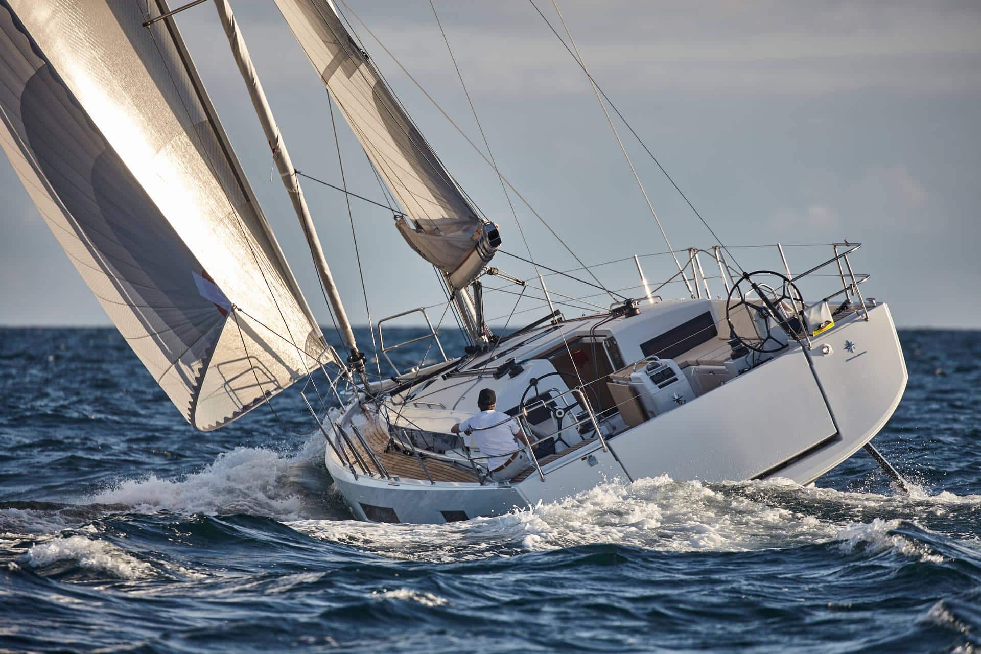 Ein Segelboot mit weißen Segeln schneidet in leicht geneigtem Winkel durch die Wellen. Eine Person steuert das gecharterte Boot, im Hintergrund sind das offene Meer und ein bewölkter Himmel zu sehen. Das Boot bewegt sich schnell und verursacht eine Gischt, wo es auf die Wellen trifft.