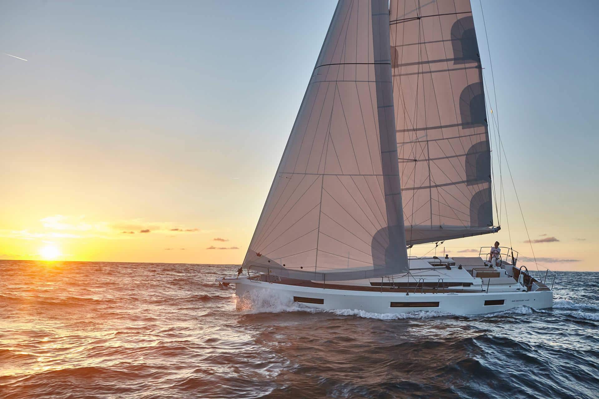 Ein Segelboot gleitet bei Sonnenuntergang mit voll ausgefahrenen Segeln über den Ozean. Die Sonne geht im Hintergrund unter und wirft einen warmen orangefarbenen Schimmer auf das Wasser, der einen Kontrast zum kühlen Blau des Meeres bildet. Der Himmel ist bis auf ein paar vereinzelte Wolken klar – die perfekte Kulisse für ein traumhaftes Yachtcharter-Abenteuer.