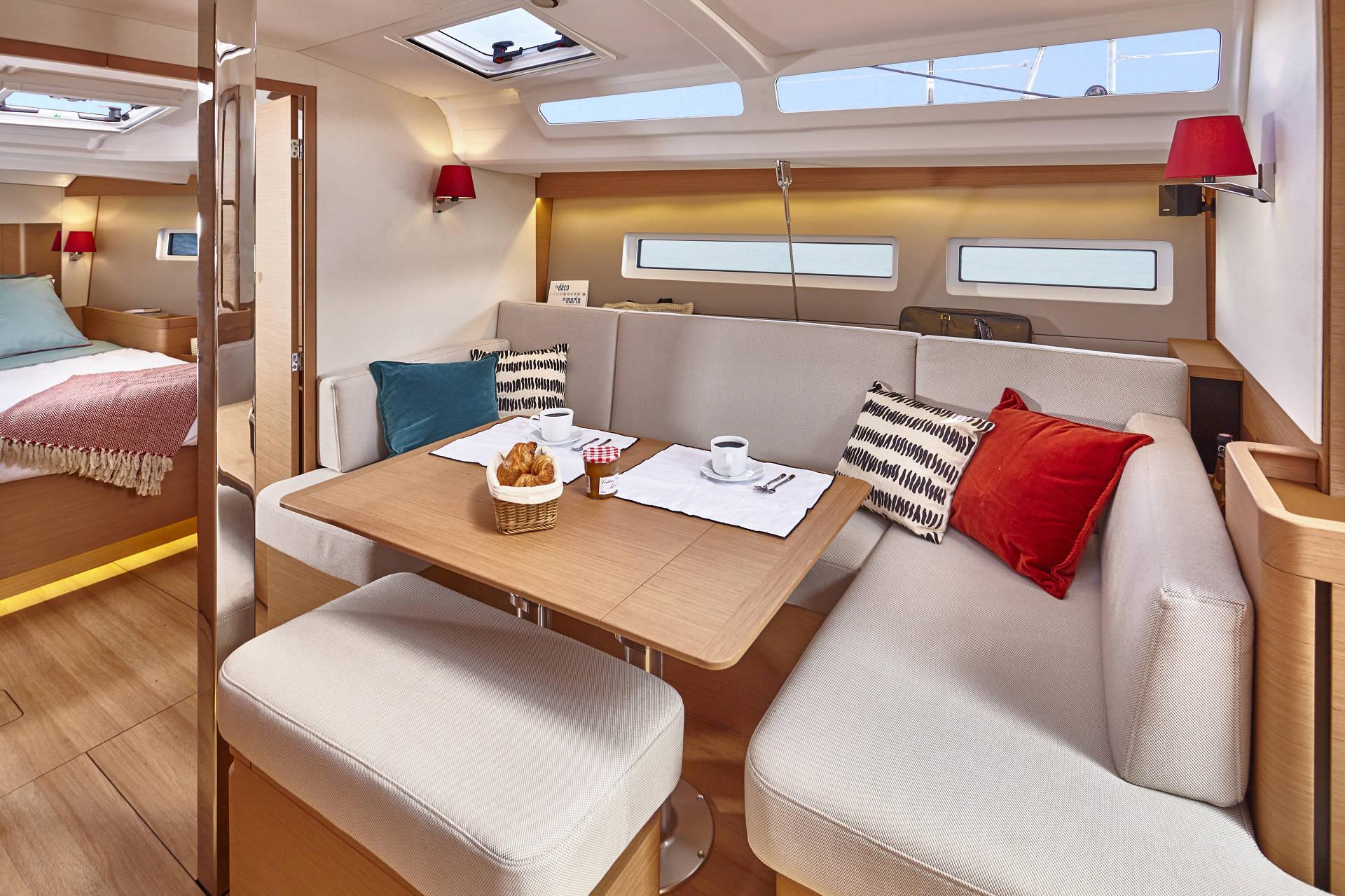 Ein gemütlicher Yachtcharter-Innenraum mit einem Essbereich mit einem Holztisch und gepolsterten Sitzgelegenheiten, akzentuiert durch bunte Kissen. Auf dem Tisch stehen zwei Gedecke mit Kaffeetassen und Croissants. Der Bereich wird durch an der Wand montierte rote Lampen beleuchtet und im Hintergrund ist ein Schlafzimmer sichtbar.