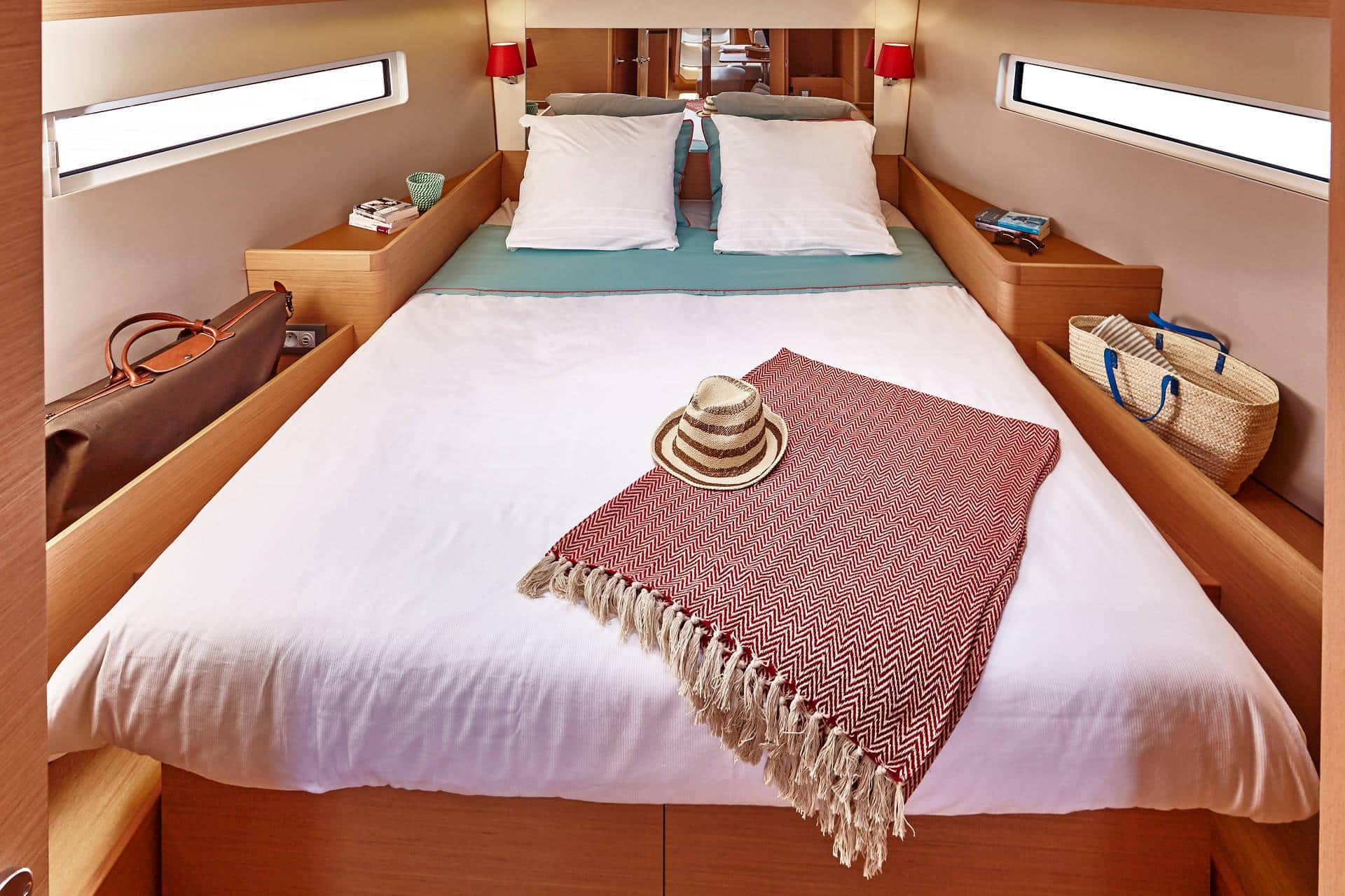 Ein gemütliches Schlafzimmer auf einem Yachtcharter mit einem ordentlich gemachten Bett mit weißer Bettwäsche und einer blaugrünen Überwurfdecke. Auf dem Bett liegt eine rot-weiß gemusterte Decke mit einem Strohhut. Das Zimmer verfügt über Holzmöbel, kleine Fenster, eine Reisetasche und einen Korb.