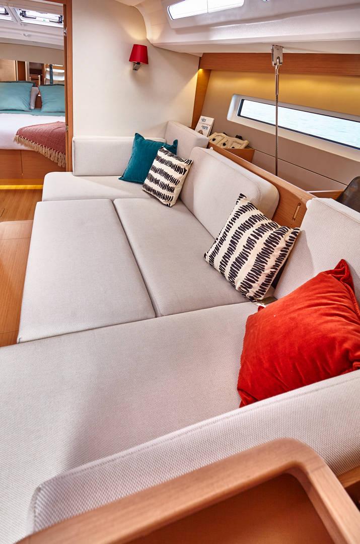 Das Bild zeigt das Innere eines modernen Yachtcharters mit einem geräumigen, hellgrauen L-förmigen Sofa, das mit bunten Kissen in Rot, Blaugrün und Schwarz-Weiß-Muster geschmückt ist. Im Hintergrund ist durch eine offene Tür ein Bett sichtbar. Der Raum ist gut beleuchtet und einladend.