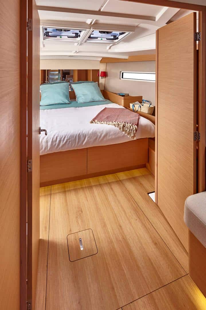 Blick auf die Kabine eines Yachtcharterboots mit einem bequemen Bett mit Kissen und einer Decke im hinteren Bereich. Die Kabine ist mit Holzpaneelen ausgekleidet und hat ein kleines Fenster über einem Regal mit persönlichen Gegenständen. Die Türen sind offen und geben den Blick auf einen hellen, gepflegten Raum frei.