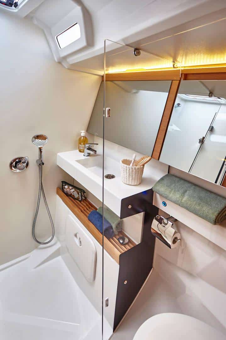 Ein modernes, kompaktes Badezimmer auf einem Yachtcharter. Es verfügt über einen an der Wand montierten Duschkopf, ein Waschbecken mit einem Spiegel darüber und Holzregale. Ein Handtuch und Toilettenartikel sind ordentlich auf den Regalen platziert. Der Raum verfügt über eine helle, effiziente Beleuchtung.