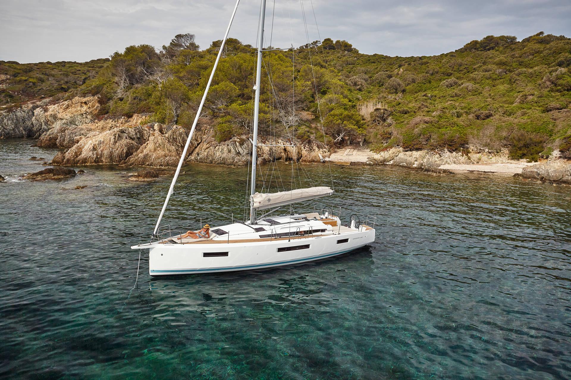 Ein elegantes, weißes Segelboot liegt in einer klaren, türkisfarbenen Bucht nahe einer üppigen, grünen, felsigen Küste vor Anker. Das Boot verfügt über ein modernes Design mit einem geräumigen Deckbereich. Perfekt für ein Yachtcharter-Abenteuer, das umgebende Wasser ist ruhig und spiegelt die grüne Landschaft und den bewölkten Himmel wider.