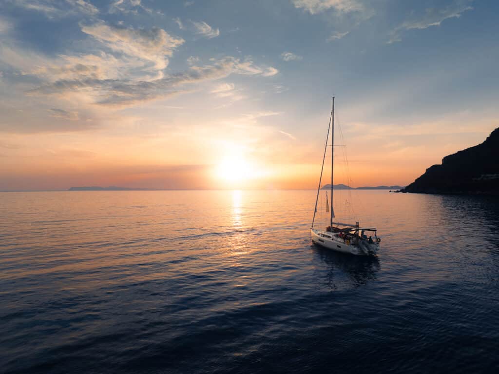 Ein einsames Segelboot gleitet bei Sonnenuntergang über ein ruhiges Meer und erinnert an eine Segelreise von Youngline. Der Himmel ist in Orange-, Rosa- und Blautönen bemalt, die sich im Wasser spiegeln. Hügelsilhouetten rahmen den Horizont auf der rechten Seite des Bildes ein und schaffen eine ruhige und malerische Szene.