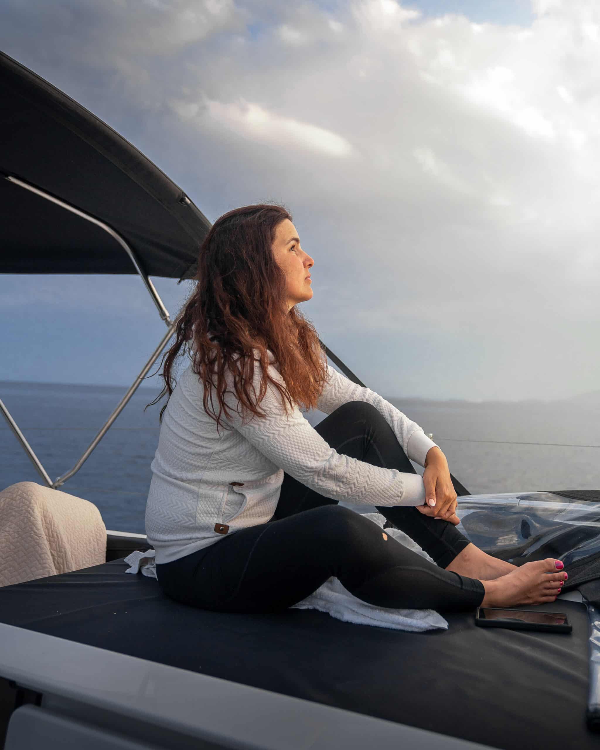 Eine Frau sitzt auf dem Deck eines Bootes und betrachtet schweigend den Himmel, während sie ihr Segelabenteuer genießt. Sie hat langes, welliges Haar und trägt einen weißen Pullover und eine schwarze Hose. Im Hintergrund sind das Meer und ein bewölkter Himmel zu sehen.