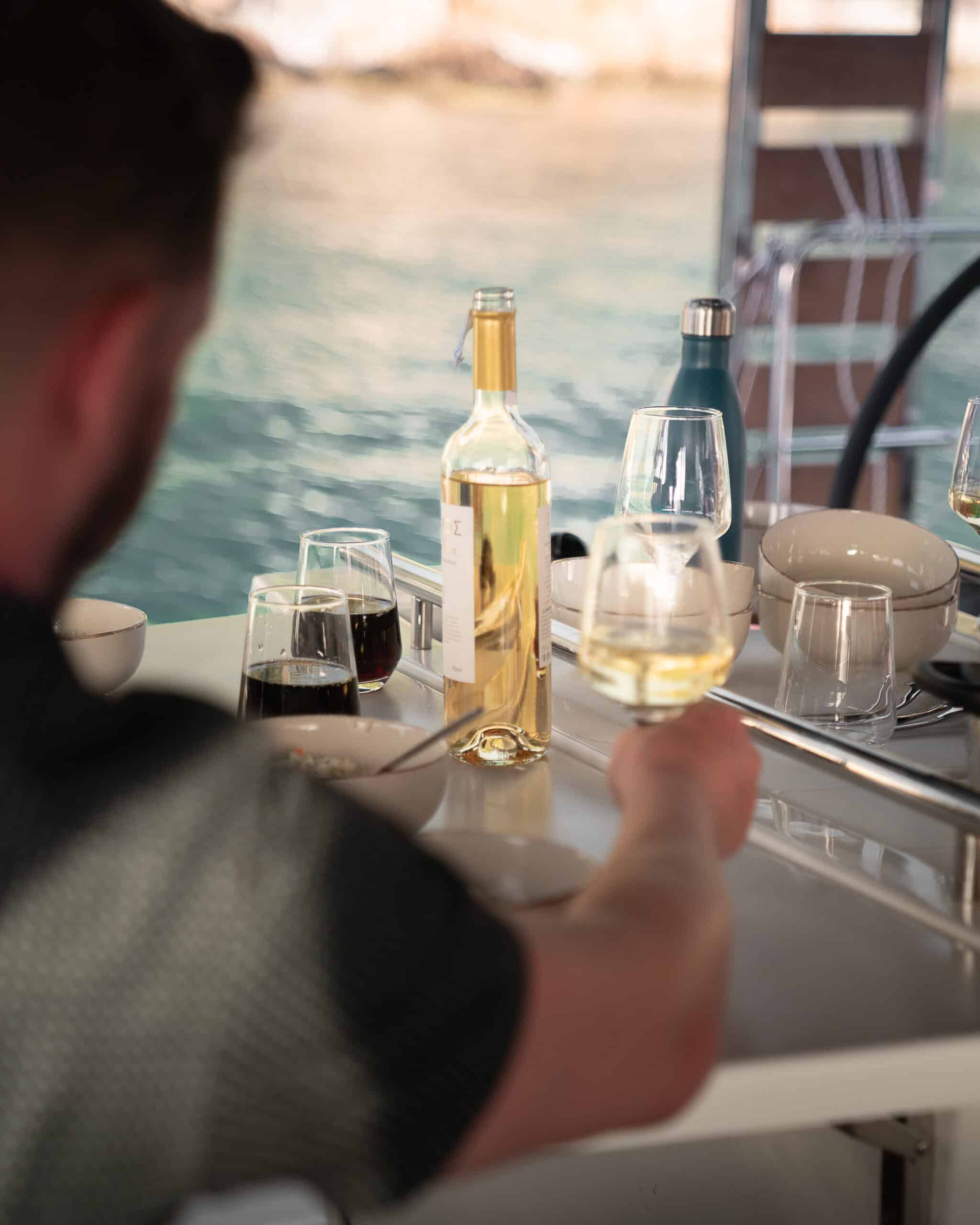 Eine Person sitzt an einem Tisch mit drei Gläsern Wein, einer Flasche Weißwein und einer Wasserflasche. Der Tisch steht vor der Kulisse eines Gewässers, was an ein Essen im Freien oder ein Segelerlebnis erinnert. Die Person greift nach einem Glas Weißwein.