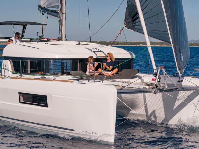 Ein moderner weißer Katamaran namens „Lagoon“ segelt an einem sonnigen Tag. Zwei Personen entspannen sich und unterhalten sich auf dem Deck vorne. Das Boot, ein Teil der Youngline-Katamaran-Flotte, gleitet durch ruhiges blaues Wasser, und andere sind am Steuer zu sehen. Im Hintergrund ist eine entfernte Küstenlinie zu sehen.