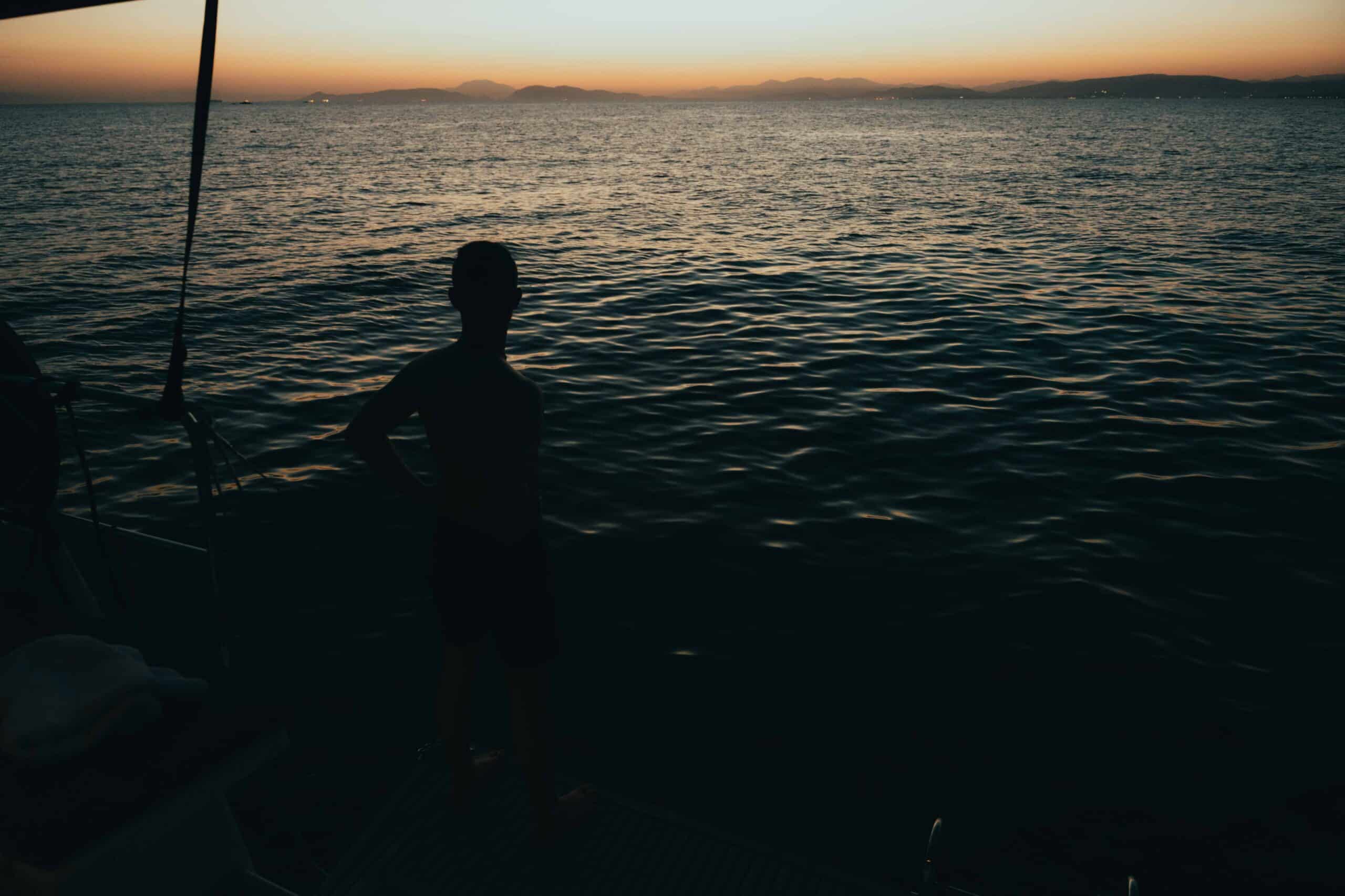Die Silhouette einer Person steht am Rand eines Bootes und blickt bei Sonnenuntergang auf ein ruhiges Gewässer. Der Himmel zeigt einen Farbverlauf von Orange zu Dunkelblau, während die Sonne hinter den fernen Bergen untergeht. Diese Segeln-Szene ruft ein Gefühl der Ruhe und Besinnung hervor.