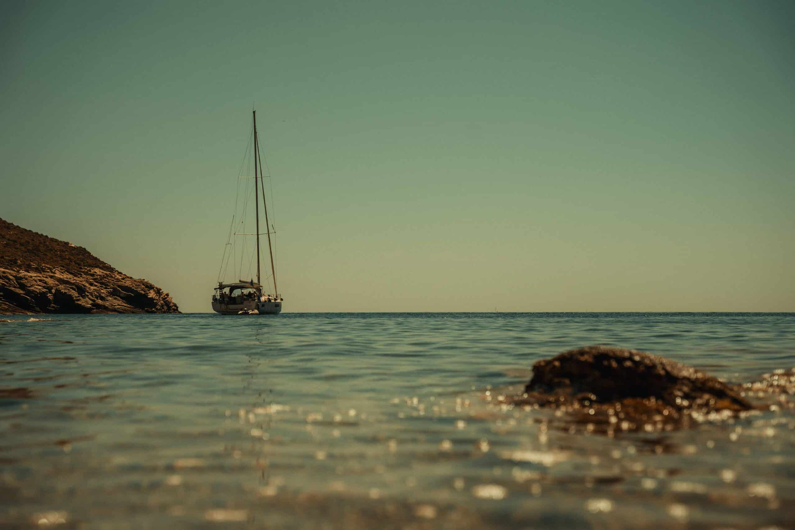 Ein Segelboot liegt in Ufernähe vor Anker. Im Hintergrund ist eine felsige Küste zu sehen. Das ruhige, klare Wasser unter einem ruhigen Himmel vermittelt ein Gefühl der Ruhe und fängt die Ruhe der Segeln-Szene ein. Im Vordergrund trägt ein kleiner, sonnenbeschienener Felsen zur malerischen Kulisse bei.
