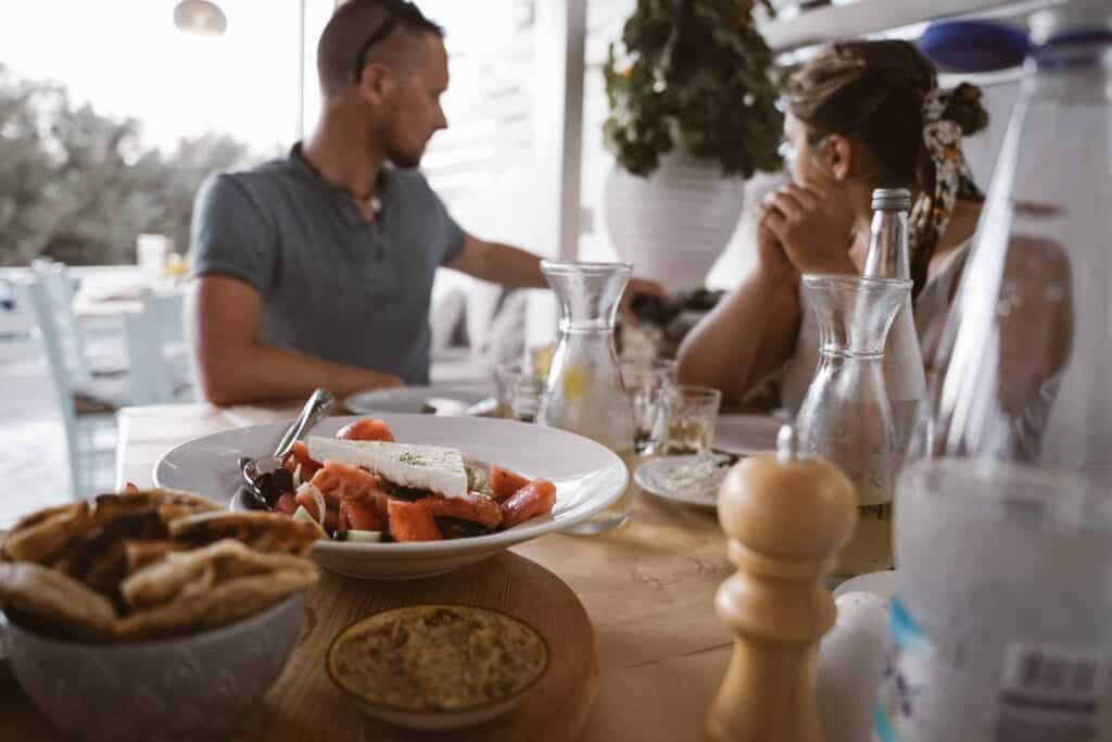 Ein Mann und eine Frau sitzen an einem Tisch, der mit verschiedenen Speisen geschmückt ist, darunter ein Salat mit Fetakäse, eine Schüssel Pitabrot und Glasflaschen. Im Hintergrund sind dekorative Gegenstände und eine Pflanze zu sehen, die eine zwanglose Atmosphäre im Freien suggerieren, die an die Kykladeninseln erinnert.