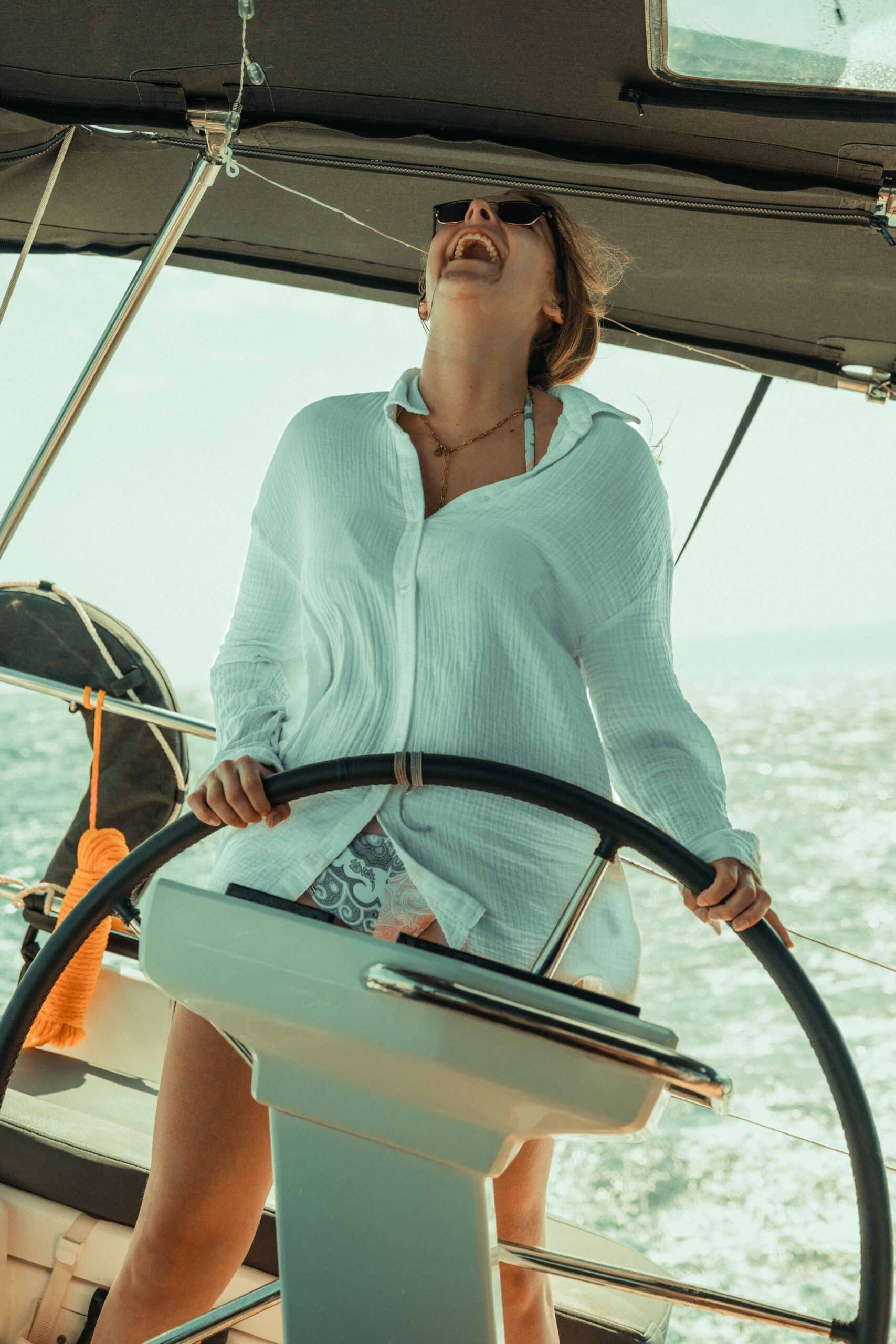 Eine Person mit weißem Hemd und Sonnenbrille steuert an einem sonnigen Tag fröhlich ein Boot. Die Person steht am Steuer, im Hintergrund ist die Meereslandschaft zu sehen, die den berauschenden Geist des Segelns verkörpert.