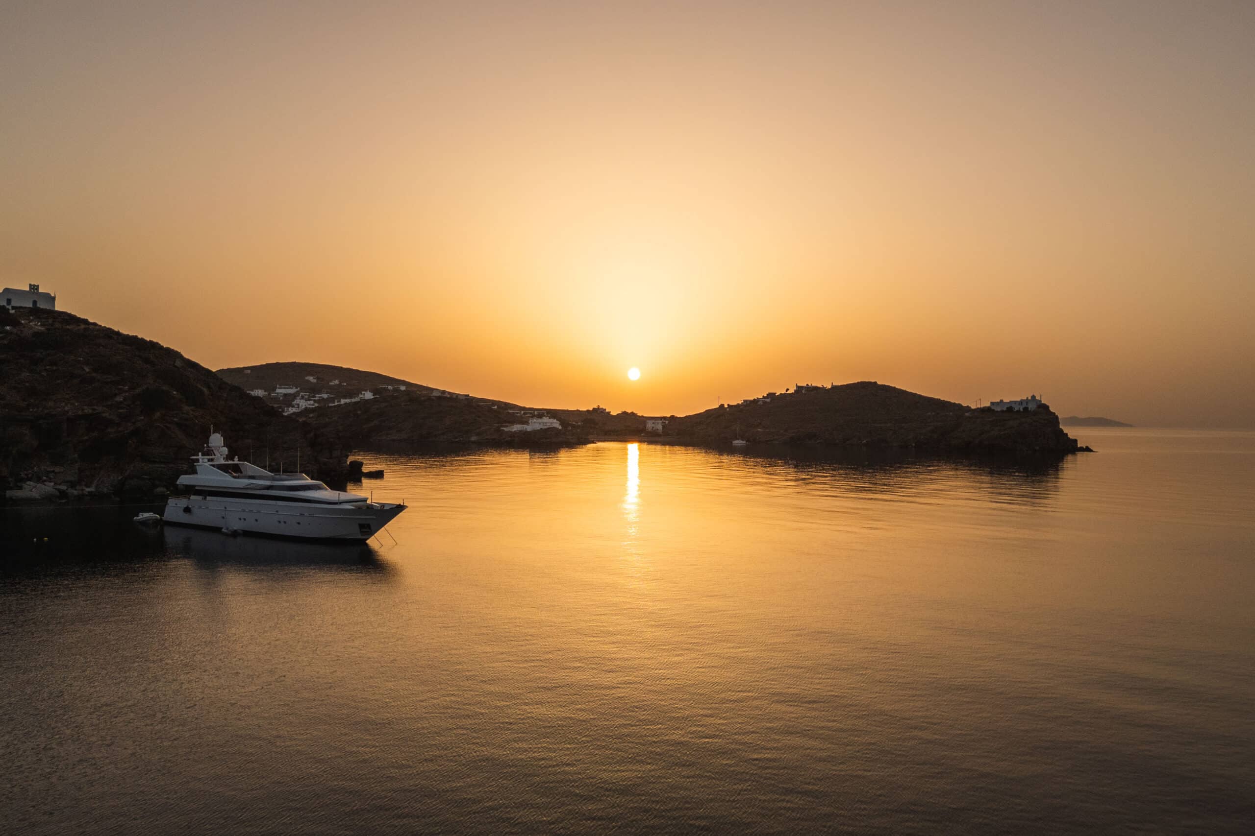 Eine große Yacht liegt bei Sonnenuntergang in einer ruhigen Bucht vor Anker – perfekt für Segelfreunde. Die Sonne geht zwischen zwei Hügeln unter und wirft einen warmen, goldenen Schimmer auf das ruhige Wasser. An den Hängen sind kleine Gebäude zu sehen, während der Himmel von einem goldenen Farbton zu Hellblau wechselt.