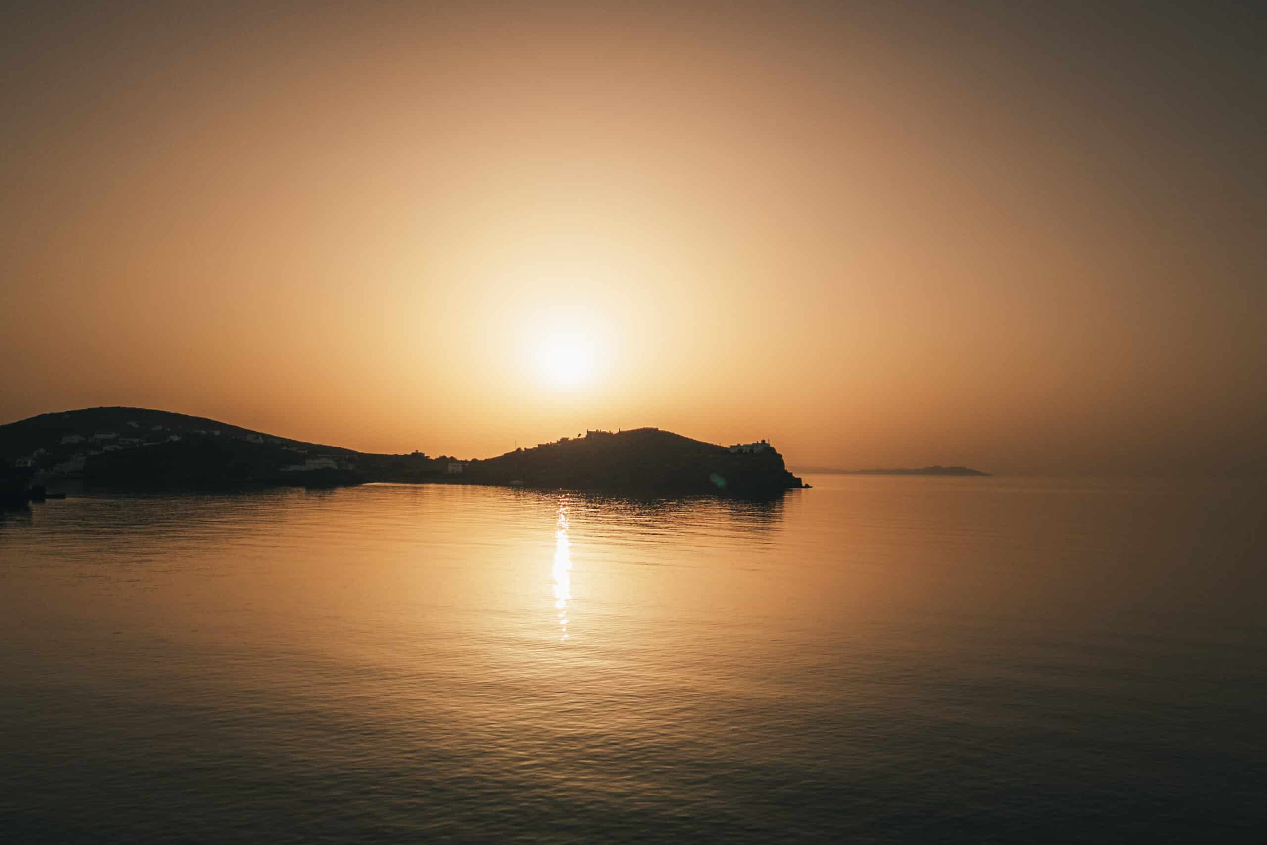Ein ruhiger Sonnenuntergang über einem ruhigen Gewässer mit klarem Himmel, perfekt zum Segeln. Die Sonne, nahe dem Horizont, wirft ein warmes, goldenes Spiegelbild auf das Wasser. In der Ferne sind Hügelsilhouetten zu sehen. Die gesamte Szene ist friedlich und ruhig.